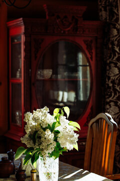 Rural interior of kitchen with bouquet of fresh white lilac flowers in glass vase on table near small brown clay pot in front of vintage red cupboard in wooden house on sunny spring day. Rural life.