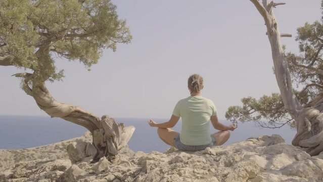 Woman meditates in lotus position while sitting facing sea on top of mountain near bonsai juniper tree.