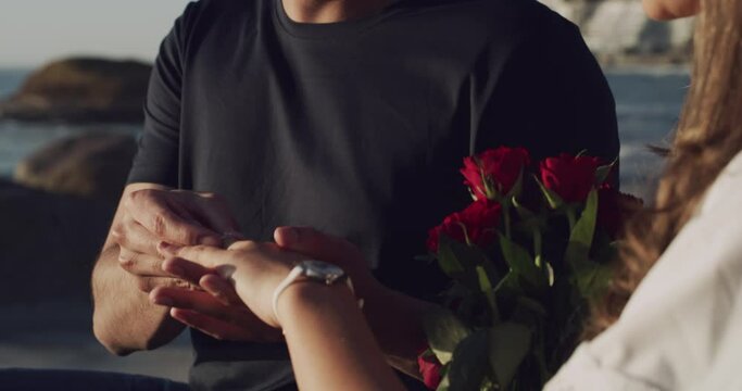 A romantic couple gets engaged after a proposal at the beach. Young woman holding a bunch of red roses showing affection to her fiance after a loving gesture as he puts a diamond ring on her finger