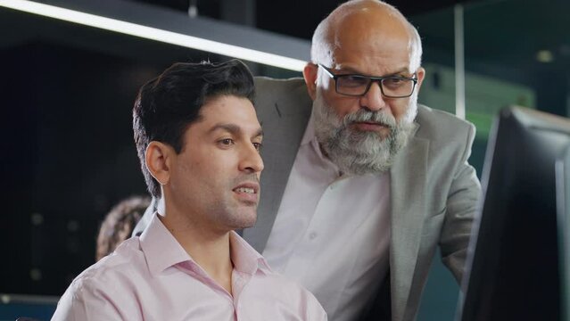 A modern Indian Asian attractive smiling customer support care male or man employee wearing headset mic working on a computer while interacting or talking to senior boss. Business, Finance concept