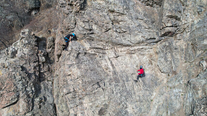 Rock climbing training on a steep slope in the mountains. The view from the drone. A group of people climb to the top of cliff. Brave girls and boys are looking for a trail on a cliff. Extreme sports