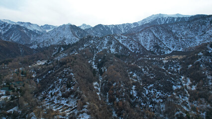 High snowy peaks stand in the distance. Drone view of the highway with cars, forest trails, blue sky with clouds. In places there is snow, a river runs. Mountainous area near Medeo, Almaty, Kazakhstan