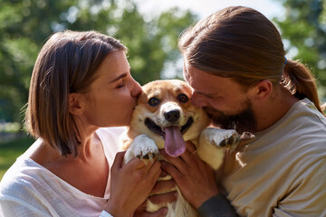 Young caucasian couple kissing and caressing adorable Corgi dog in park.