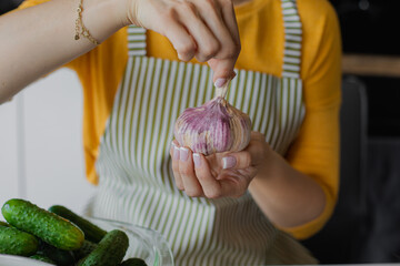Closeup unrecognizable woman in apron holding garlic bulb near fresh cucumber plate in kitchen. Immunity and healthcare