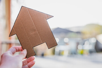 hand holding house icon in front of house exterior and backyard with table an chairs and mountains view, concept of real estate or dream home or family life