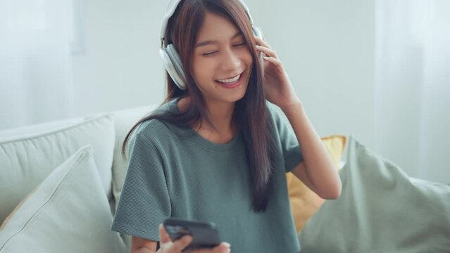 Happy young asian woman wear wireless headphones having fun listen music in smartphone and sit on sofa in living room, Female enjoys listening chill music audio sound feeling no stress at home.