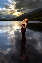 Fototapeta premium Woman with long red hair standing in the still water of a lake with a dramatic cloudy sunset