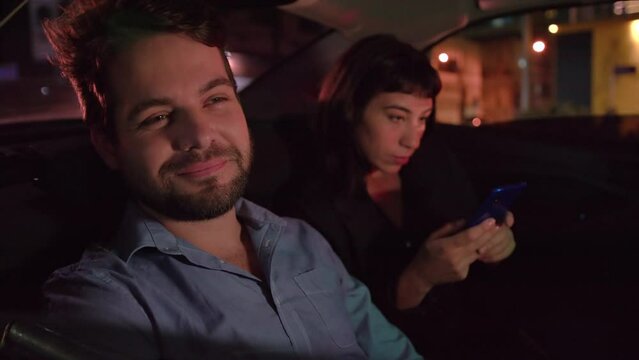 Happy man commuting from work in taxi car backseat at night looking out vehicle window. Couple inside cap