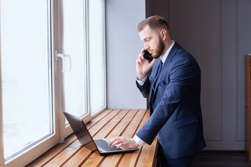 Young businessman in formal wear talking on the phone and analyzing data using laptop in the office