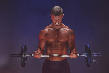 Fototapeta na wymiar Brutal sweaty strong young man athlete with naked upper body standing doing workout with barbell and showing strong pumped up biceps over smoky background. Sport men body concept