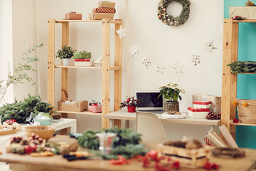 Interior of modern workshop full of Christmas decorations on workspaces: Christmas wreath on wall, star garland hanging between shelves with gift boxes