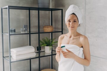 Attractive caucasian girl wrapped in towel after bathing and hair washing is holding a cream jar.