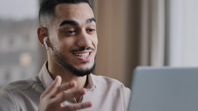 Smiling hispanic man arab guy wears wireless earphones using electronic device talks at computer web camera makes call video conference remote job interview greets says hello consulting client online 
