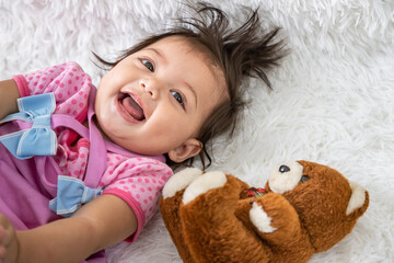 Little Asian baby girl lying down on carpet at home with teddy bear doll. Newborn child relaxing in bed. Nursery for young children.