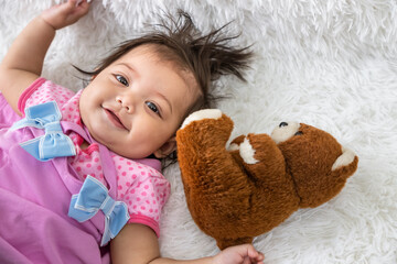 Little Asian baby girl lying down on carpet at home with teddy bear doll. Newborn child relaxing in bed. Nursery for young children. Looking at camera.