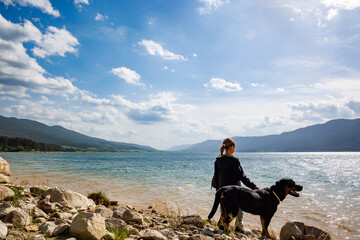 Girl stands near dog of Rottweiler breed on shore near lake against backdrop of mountain range covered with forest