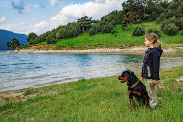 Girl stands near dog of Rottweiler breed in meadow next to lake, against hilly valley with spruce forests