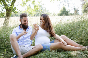 young couple in love sitting in high flower meadow in summer and cuddling and the woman is pregnant and the man has a full beard