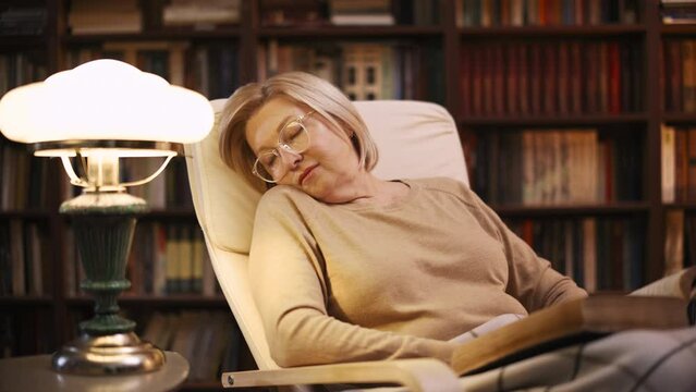 Senior woman falling asleep while reading a book in her library, late evening