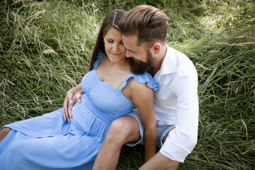 young couple in love sitting in high flower meadow in summer and cuddling and the woman is pregnant and the man has a full beard