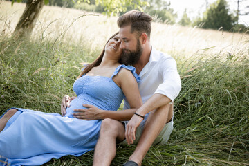Naklejka na ściany i meble young couple in love sitting in high flower meadow in summer and cuddling and the woman is pregnant and the man has a full beard