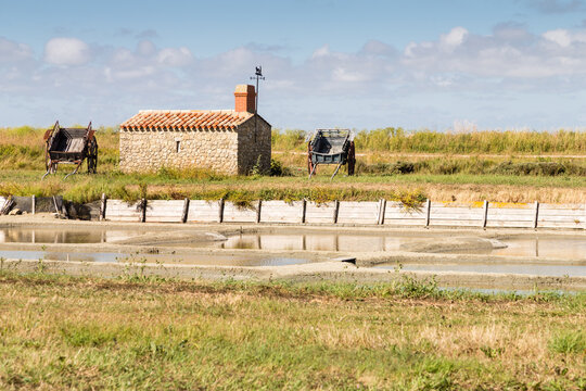 Petite maison en pierres avec girouette et deux charrettes en bois devant un marais salant. Marais des Ileaux, Noirmoutier