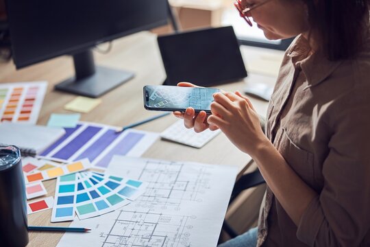 Woman taking a photo of a blueprint and color samples palette using smartphone while working on new interior design project in her office