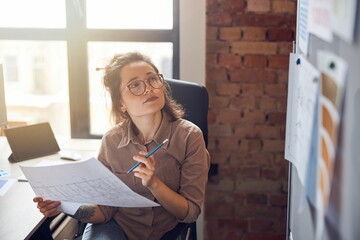 Enthusiastic young woman designer holding blueprint paper looking at board while working on new interior design project in her office