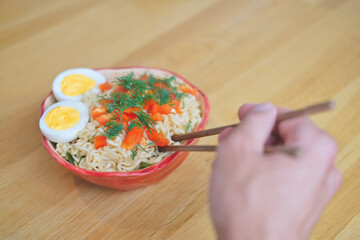 man (hand) eating asian egg noodles in red bowl with bamboo chopsticks close up