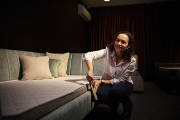 Obraz premium Charming female interior designer, seller in furniture store cleans the dust of a comfortable sofa bed with orthopedic mattress, looking at camera