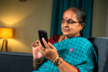Senior woman busy using mobile phone while sitting on sofa at home - concepts of technology,...