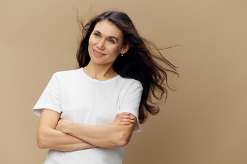 Fototapeta premium pleasant, funny woman with long well-groomed hair flying in the wind stands in a white T-shirt with her arms folded on her chest and smiles broadly with beautiful bright teeth with her eyes closed