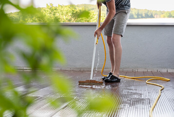 Body of a man with a scrubbing brush and a water hose making spring cleaning on a wooden terrace