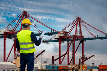 A port worker in a yellow vest with drawings in his hands against the background of port cranes and sea containers.