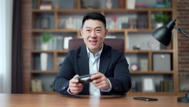Asian man in suit in office at work playing computer games on console joystick, sitting at table desktop, smiling, happy, relaxed. Cheerful business man during a break in free time videogames