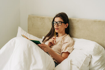 Smart student girl reads a book in bed and makes notes with a pencil.