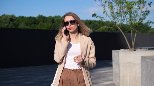 Successful pushful business woman speaking on mobile phone in park area. Businesswoman stands outdoors talking by mobile phone answering business call. Female professional having phone conversation.