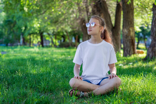 Teenage girl in white tee basic youth apparel shoot. A girl in sunglasses sits on the grass in a city park. Solid color clothing with space for text or pattern. 