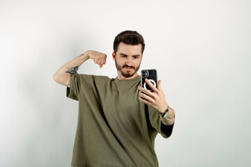 Handsome young man wearing casual clothes posing isolated over white background taking selfie, showing strong arm, flexing biceps and posing for photo.