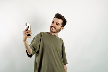 Happy young man wearing khaki t-shirt posing isolated over white background making a selfie looking at the phone screen with big smiles.