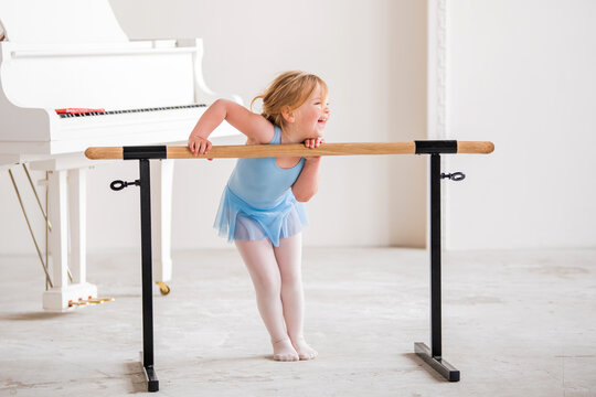 cute plump ballerina in a blue leotard is doing ballet dancing at the ballet barre in a large bright classroom.