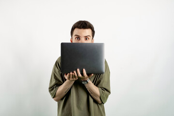 Caucasian young man wearing t-shirt posing isolated over white background peeking out from behind laptop, looking surprised at camera.