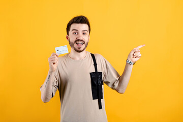 Cheerful young man wearing t-shirt posing isolated over yellow background holding credit bank card and pointing index finger aside. Shopping and finance concept.