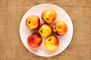 Several ripe juicy nectarines with a white ceramic plate on jute fabric, close-up, top view.
