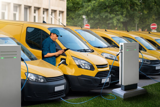 A man with a digital tablet stands next to yellow electric delivery vans at electric vehicle charging stations