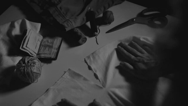 Woman leans a piece of cloth on a table. A seamstress is about to start mending a tear. Dramatic black and white scene with tailoring items.
