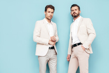 Portrait of two handsome confident stylish hipster lambersexual models. Sexy modern men dressed in white elegant suit. Fashion male posing in studio near blue wall