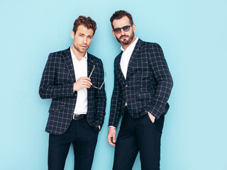 Portrait of two handsome confident stylish hipster lambersexual models. Sexy modern men dressed in black elegant suit. Fashion male posing in studio blue white wall. In sunglasses