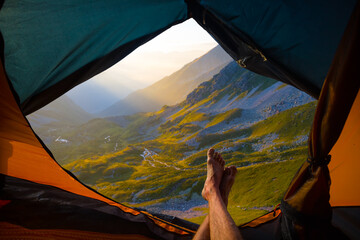 closeup hiker legs  lie in touristic tent on mountain valley background, view from touristic tent to early morning mountain valley