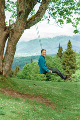 A young man on a swing against the backdrop of mountains, resting in nature, enjoying vacation, approaches, relaxation